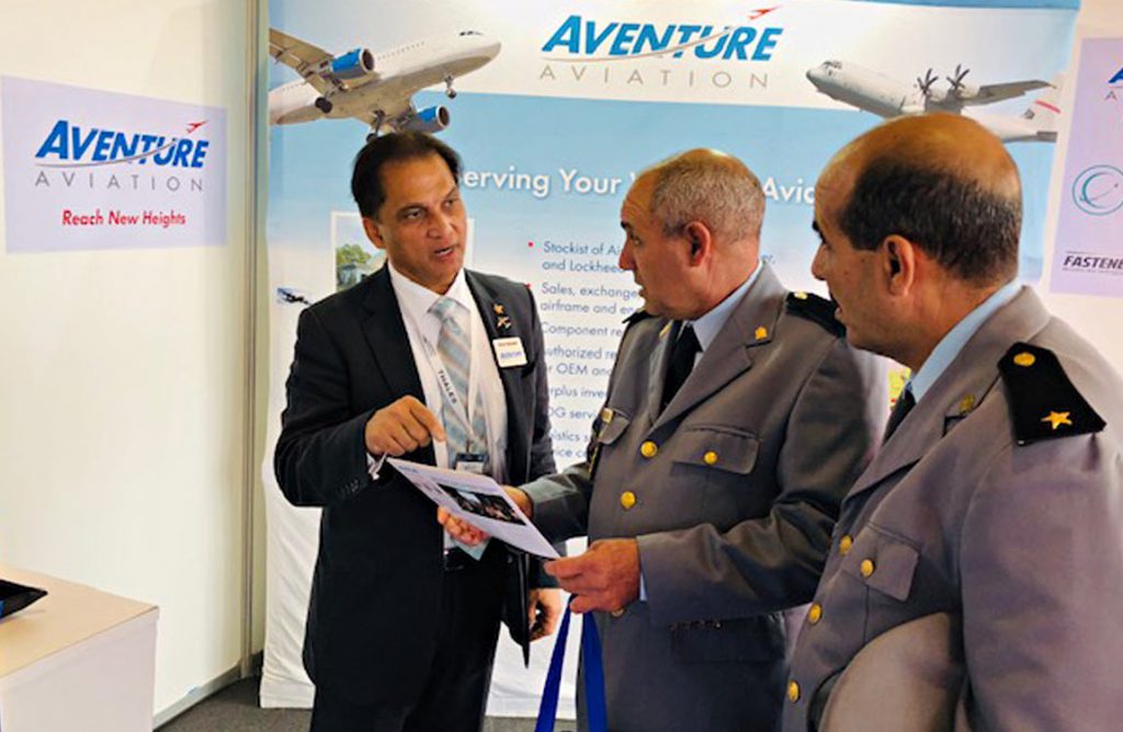 A man in a business suit shows an item to two male military officers at an Aventure Aviation trade show booth 