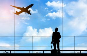 A man with carry-on baggage inside an airport looks out a huge glass window to see a commercial airliner take off among a blue sky with beautiful clouds