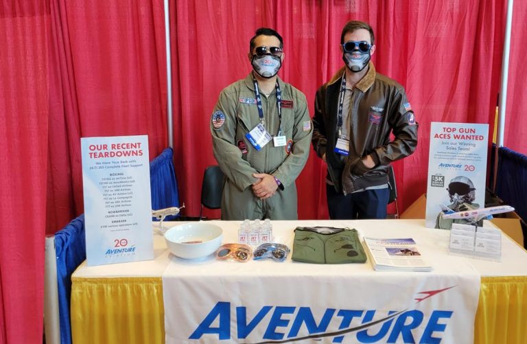 Two men dressed in jet fighter uniforms in front of exhibition booth with Aventure Aviation logo