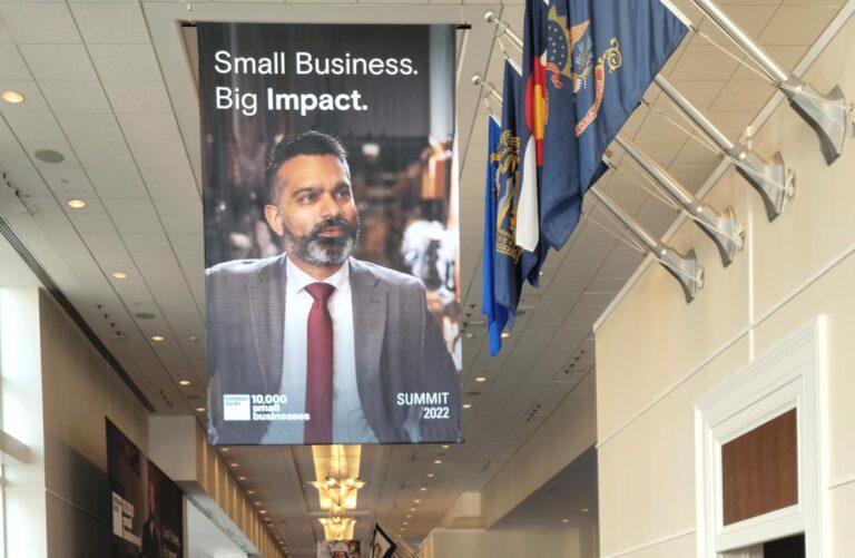 A banner hanging from the ceiling featuring Aventu re president Talha Faruqi and the words "Small Business Big Impact" on display next to U.S. flags, in a long corridor