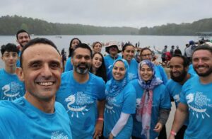 14 members of the Islamic Speakers Bureau boating team, wearing matching blue team shirts, smile in front of Lake Lanier, Georgia, with boats in distance