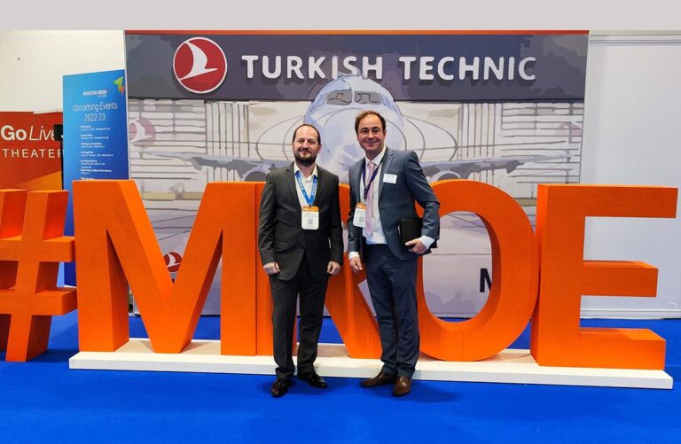 Two men in a convention hall in front of large 3D text saying "#MROE", under a sign saying "Turkish Tecfhnic"