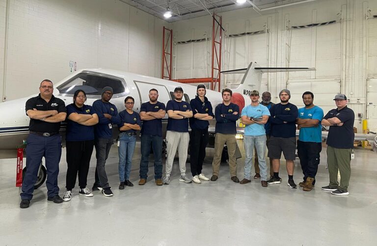 A group of 13 students and instructors stand next to each other in fromt of a small commercial airplane inside a hangar