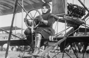 Historic photo of Blanche Stuart Scott, sitting in an early biplane on the ground, holding a large steering wheel