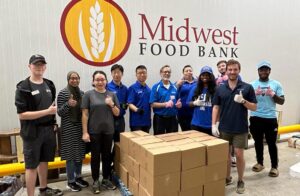 A group of 11 people smiling in front of a pile of boxes, below a large sign saying "Midwest Foodbank"