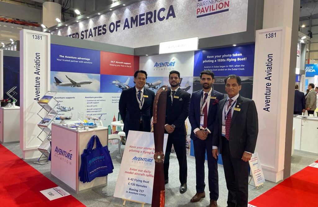 Four men stand in front of a trade show booth saying "Aventure Aviation," under a large sign saying "United States of America Pavilion"