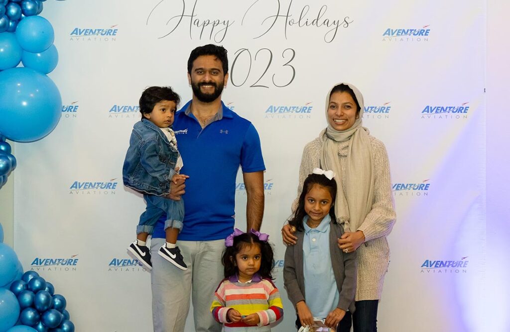 A family of five smiling in front of a "step and repeat" backdrop saying "Aventure Aviation Happy Holidays 2023 " with blue balloons