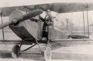 A female African American aviator (Bessie Coleman) in a long trenchcoat posed for a photo before climbing into the cockpit of a 1920s biplane