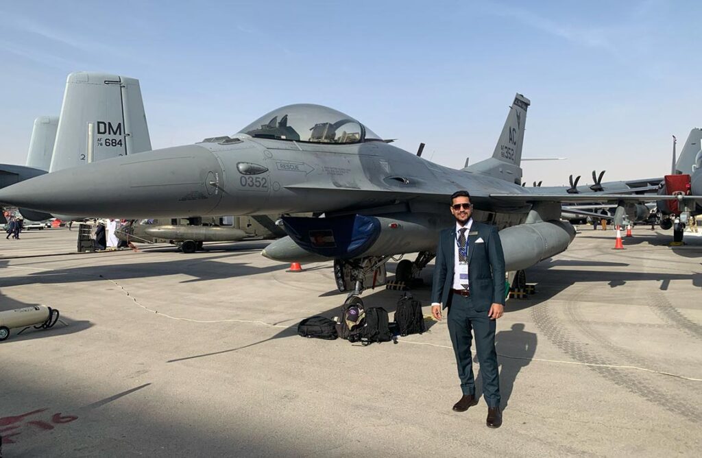 A man wearing a business suit and sunglasses posed in front of an F-16 fighter het on the ground, with other military aircraft in the background