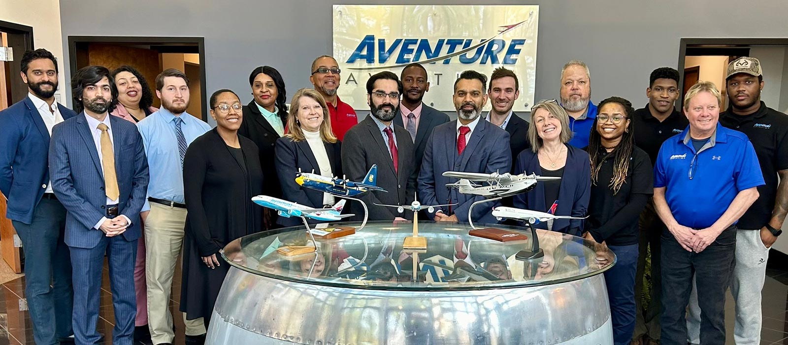 A group of 18 staff members, smiling and facing the camera, inside a building lobby with a sign saying Aventure Aviation, and a table with various model aircraft