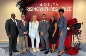 A group of six men and women in business attire posing for a photo in front of a sign with the Delta Air Lines logo and text saying "Rising with Resilience"