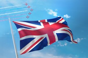 Large United Kingdom flag in front of a blue sky with six jet aircraft flying in formation small in the background.