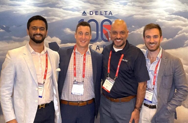 Four men smile towards camera against a backdrop saying "Delta 100 Years"