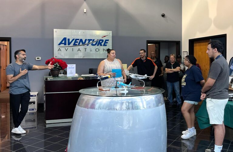 Eight people interacting in a bulding lobby, with a sign on the wall saying "Aventure Aviation" and model aircraft on a table made of an aircraft engine