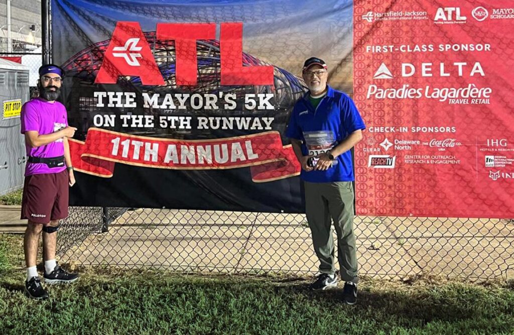 Two men dressed in running clothes standing outside, next to a sign that says "ATL - The Mayor's 5K on the 5th Runway 11th Annual