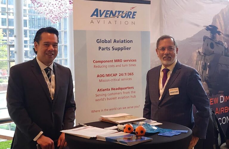 Two men stand at a table full of brochures and flyers, in front of a sign saying "Aventure Aviation – Global Aviation Parts Supplier"/