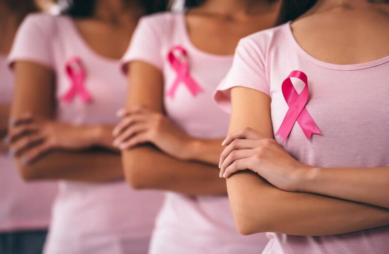 Three women, each with their arms crossed, wearing a pink shirt with a pink ribbon