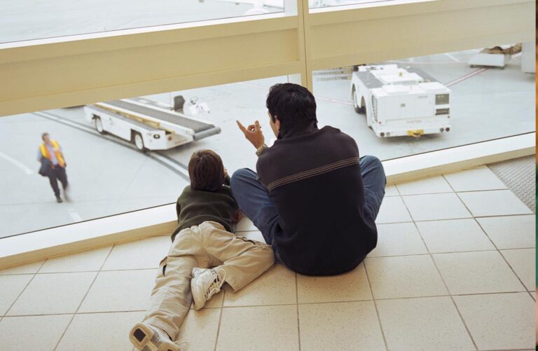 A man and child inside an airport terminal look out the window, with the man pointing out something on the tarmac