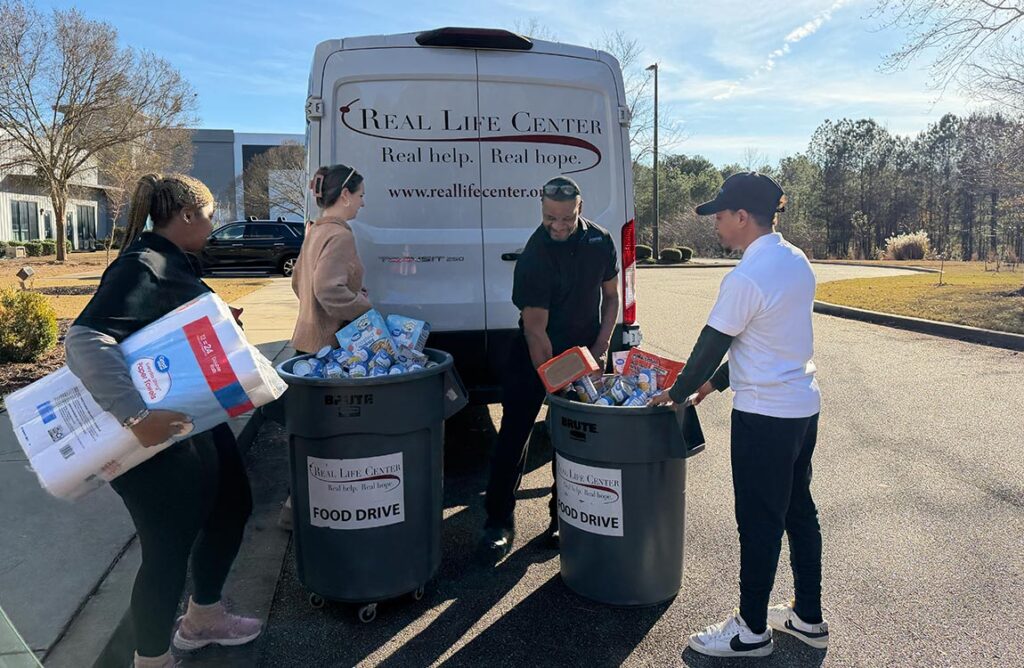 Four people prepare to load large containers of food and household goods into the back of a commercial van