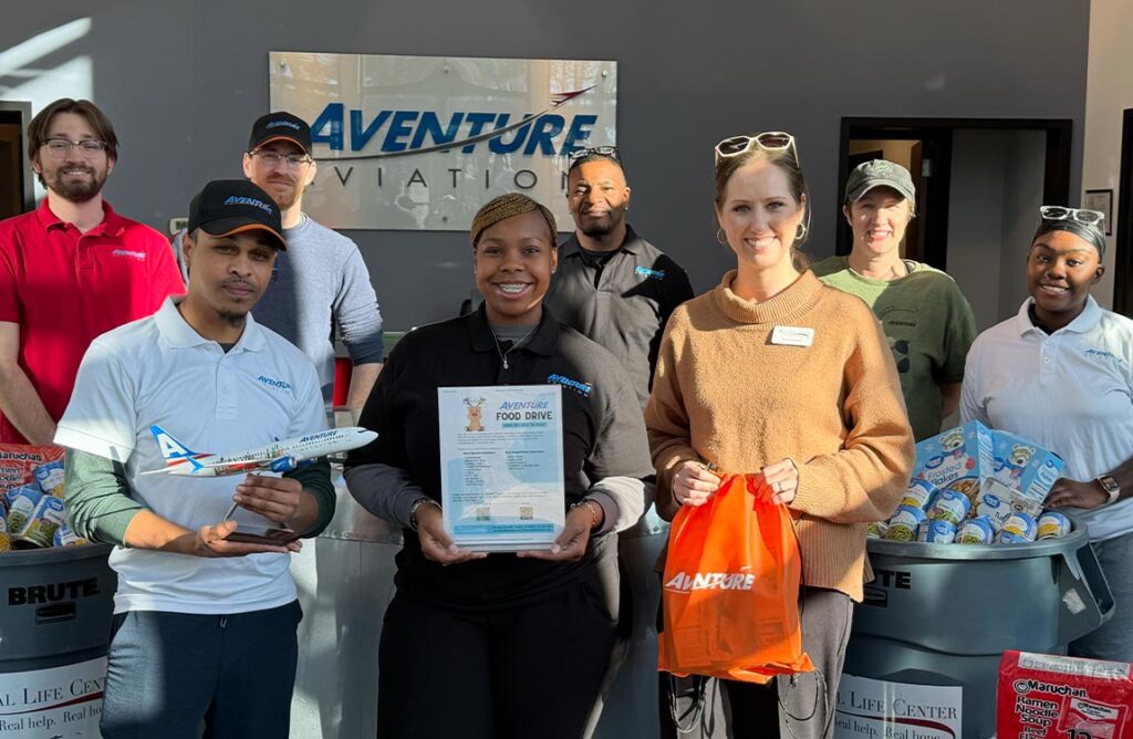 Eight people hold up donations and other items during a food drive, with a sign in the background saying "Aventure Aviation"