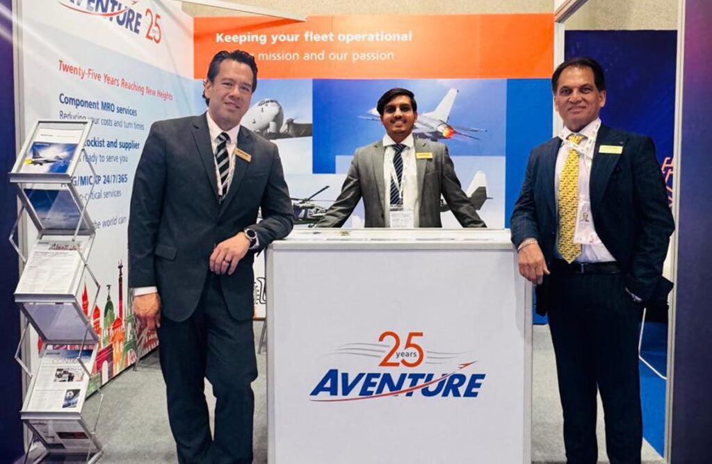 Three men in business suits stand inside a trade show booth next to a counter with a logo saying "Aventure 25 Years"