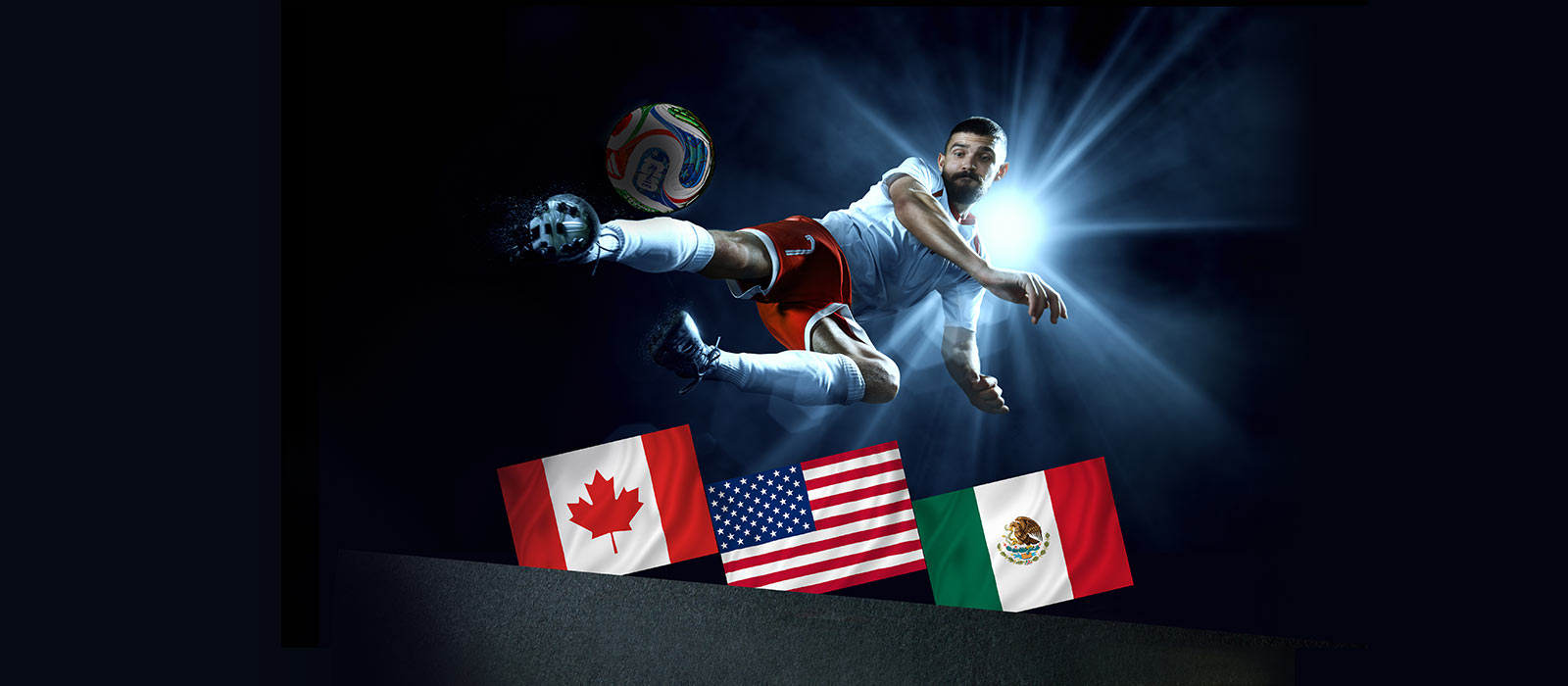 A soccer player in a stadium at night in front of a spotlight, kicks a ball in the air above the flags of Canada, USA, and Mexico