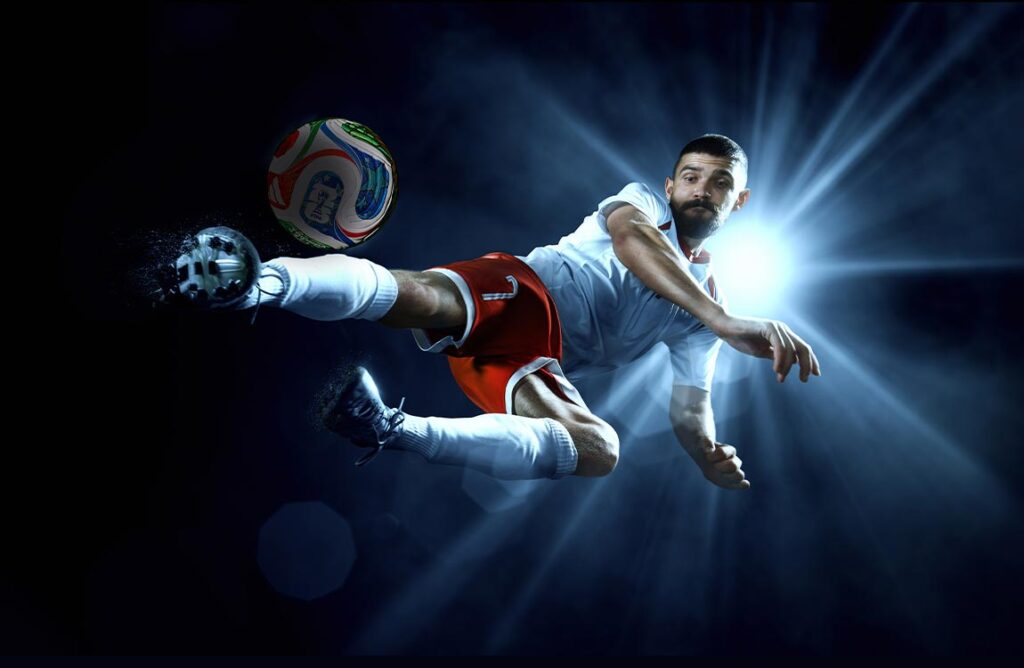 A soccer player in a stadium at night in front of a spotlight kicks a ball in the air
