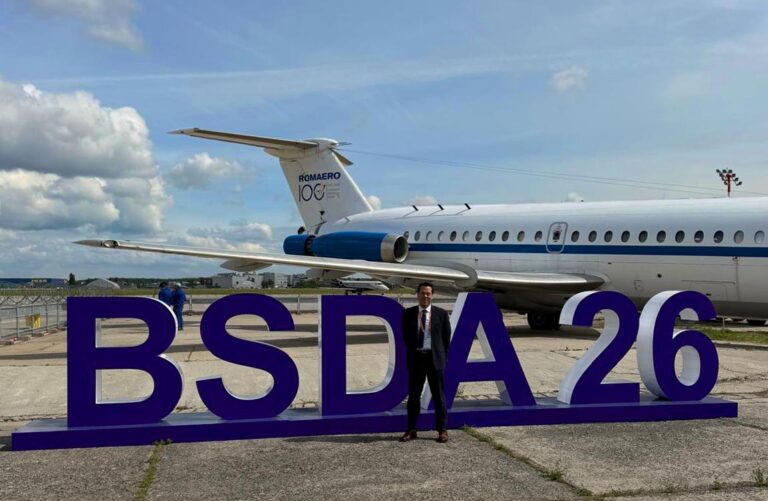 Man wearing a suit standing in front of huge letters saying BSDA26 on an airport tarmac, with a large commercial jet in the background under blue sky