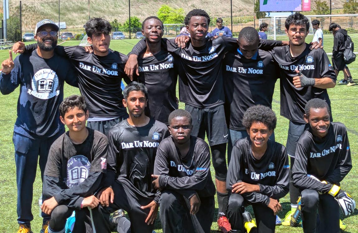 Team photo of 11 high school age boys wearing soccer shirts saying "Dar Un Noor Academy" on a soccer field