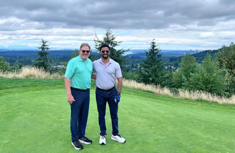 Two men with sunglasses smile on a golf course green which overlooks an expansive valley